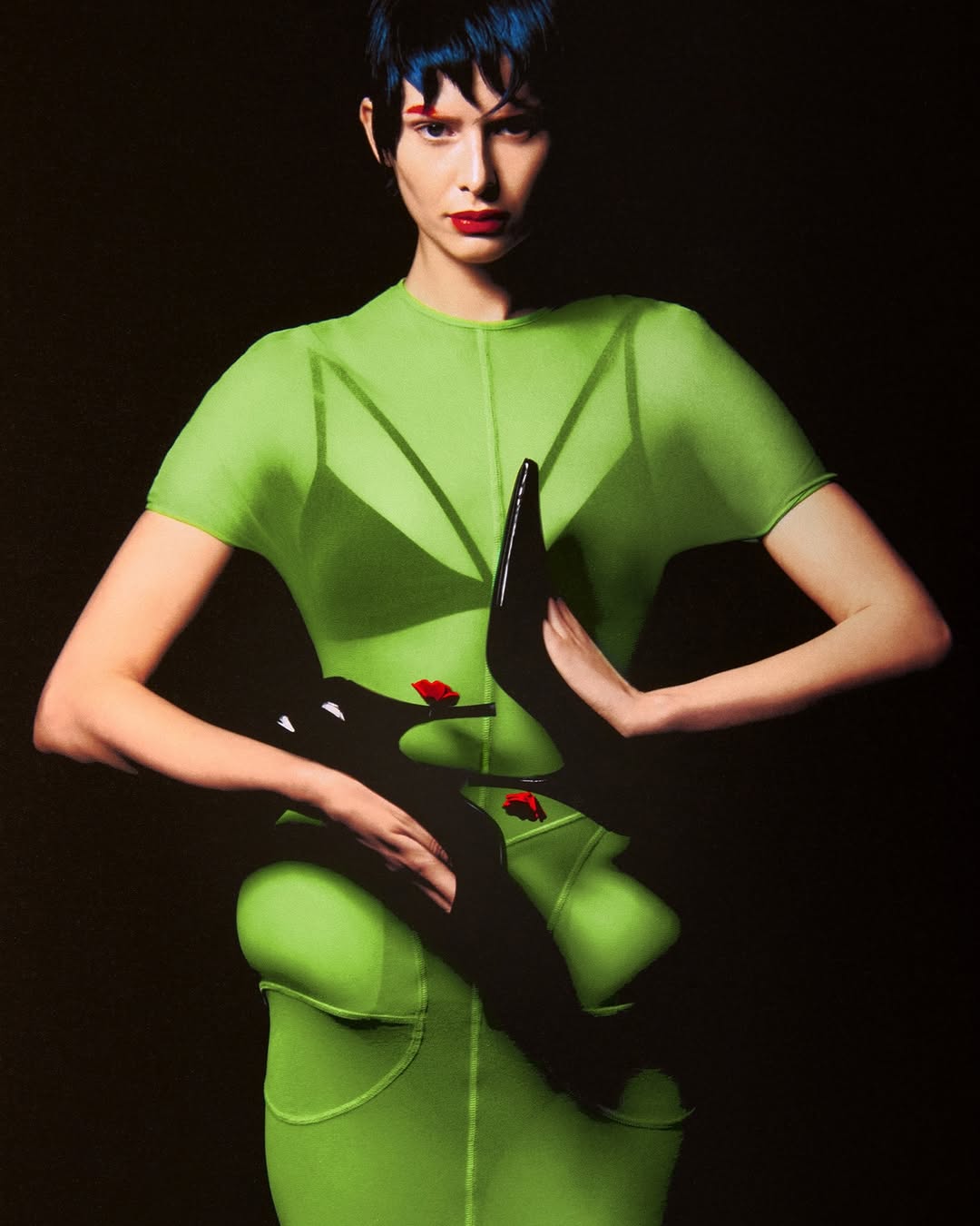 A slender woman poses in a green, semi-sheer outfit, holding a pair of black high heel shoes against a dark backdrop in a fashion photo.