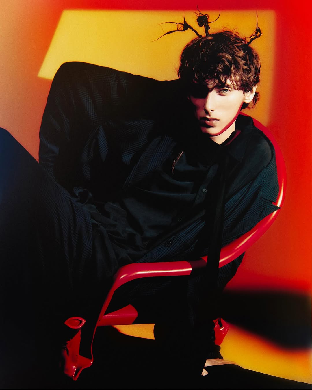 Indoor portrait of a young adult male wearing dark shirt, seated on a glossy red chair against a color block orange background.