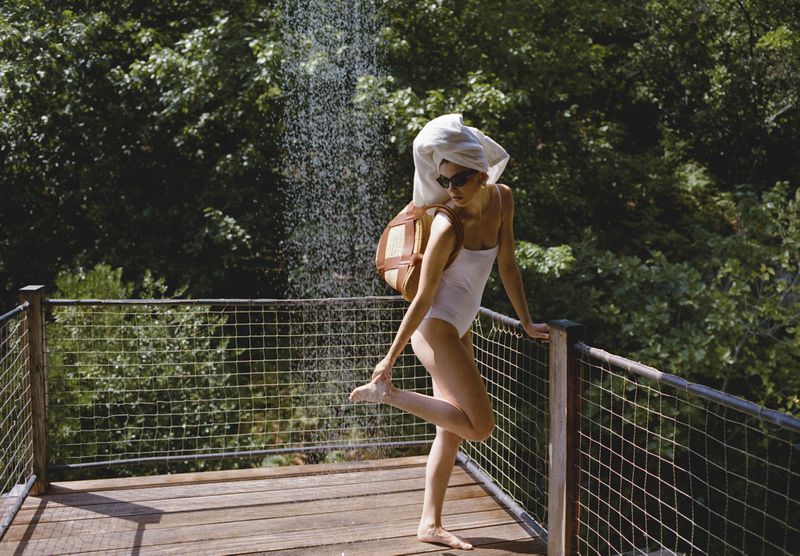 Woman in white swimsuit on wooden deck under outdoor shower with backpack and towel. Lush green trees fill background.