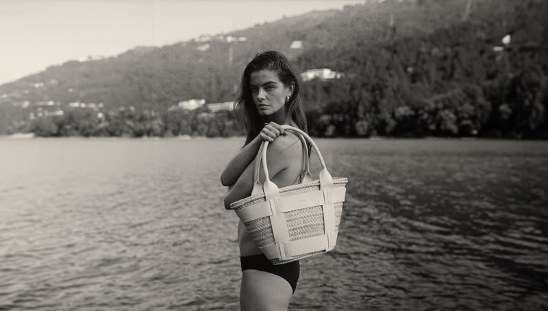Woman on a lake shore holding a straw tote bag with leather trim, with mountains in the background in a black and white photograph.