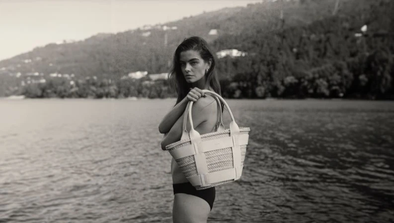 Woman on a lake shore holding a straw tote bag with leather trim, with mountains in the background in a black and white photograph.