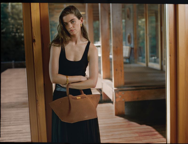 Indoor shot of a woman in a black top and skirt, holding a brown handbag, with a relaxed facial expression, against a wooden interior background.