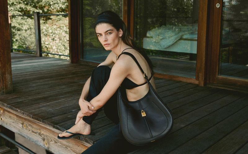 A woman in a black top and pants sits on a wooden porch with a black shoulder bag, looking towards the camera in natural lighting.