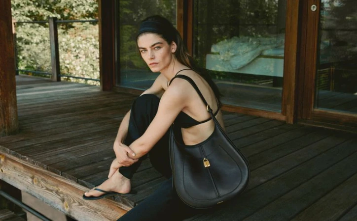 A woman in a black top and pants sits on a wooden porch with a black shoulder bag, looking towards the camera in natural lighting.