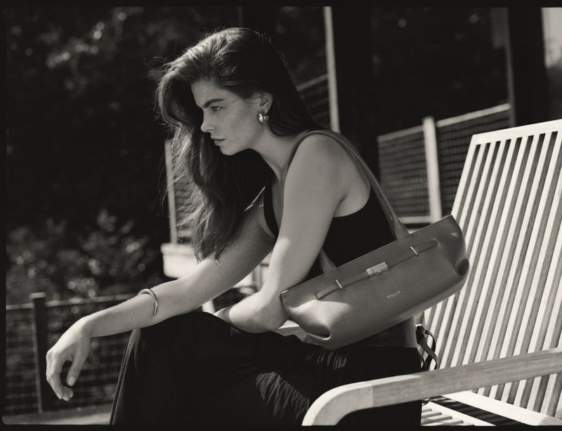 Monochrome portrait of an adult female seated on a slatted bench with a shoulder bag, wearing a tank top and bracelet.