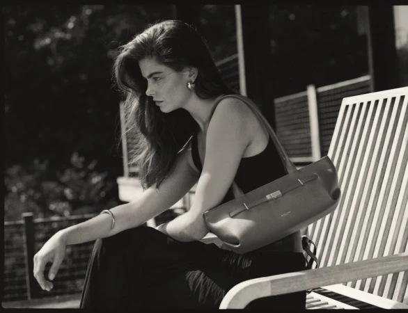 Monochrome portrait of an adult female seated on a slatted bench with a shoulder bag, wearing a tank top and bracelet.