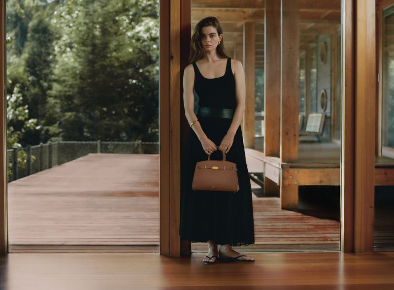 An elegant woman poses indoors wearing a black dress and holding a brown leather handbag, creating a polished fashion editorial aesthetic.