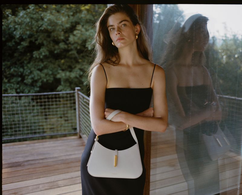 Indoor shot of a woman in a black dress, leaning against glass. She is holding a white purse. Green trees are seen in the background.