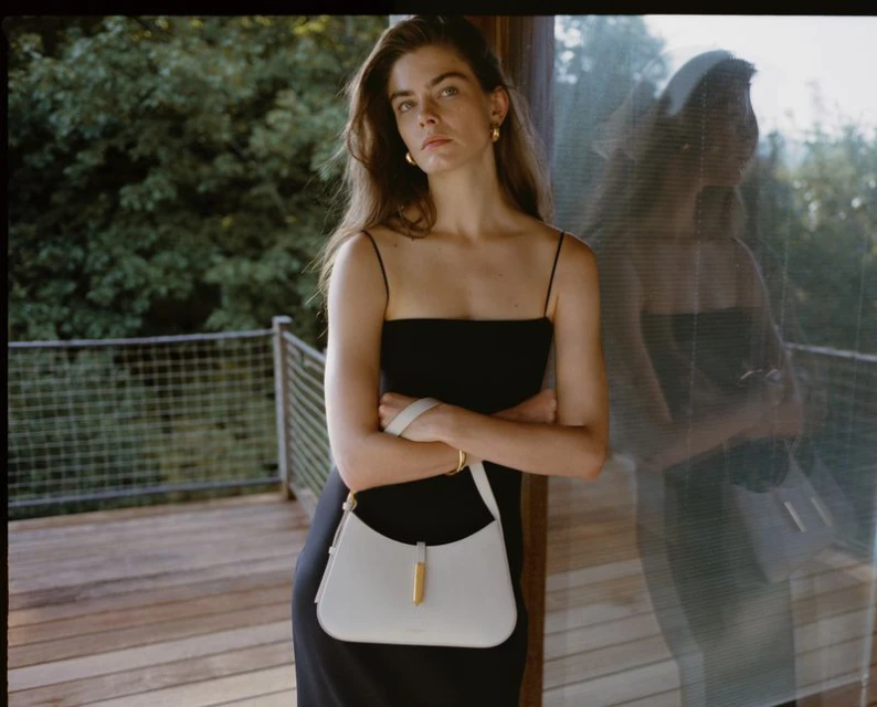 Indoor shot of a woman in a black dress, leaning against glass. She is holding a white purse. Green trees are seen in the background.