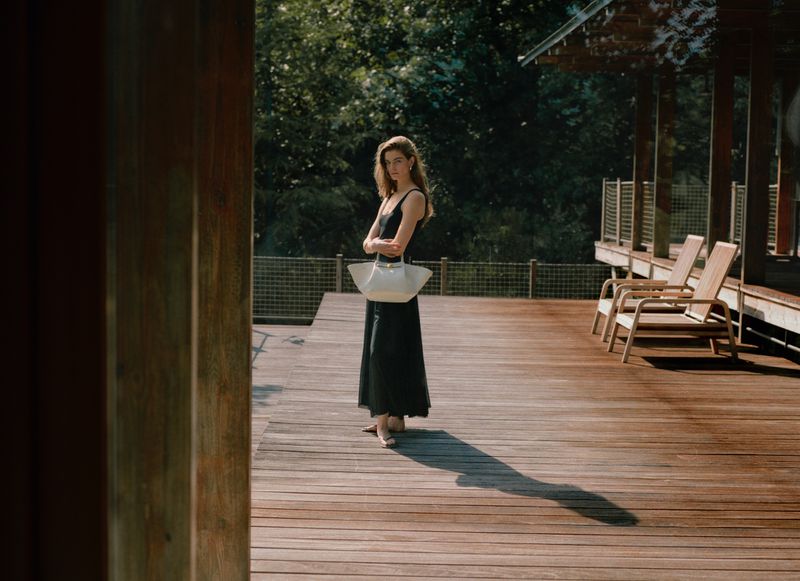 Woman in a black dress stands on a wooden deck near chairs, trees, and greenery, holding a white tote bag and glancing at the camera.