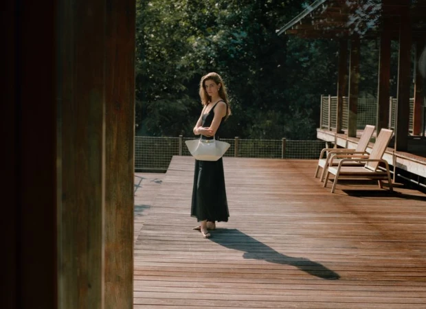 Woman in a black dress stands on a wooden deck near chairs, trees, and greenery, holding a white tote bag and glancing at the camera.