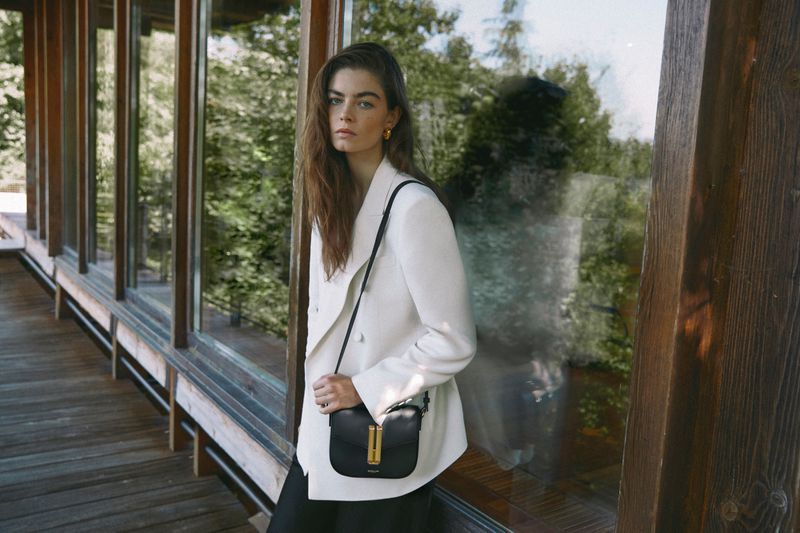 Brunette woman wearing a white double-breasted blazer and carrying a black bag stands near a window with a green tree view.