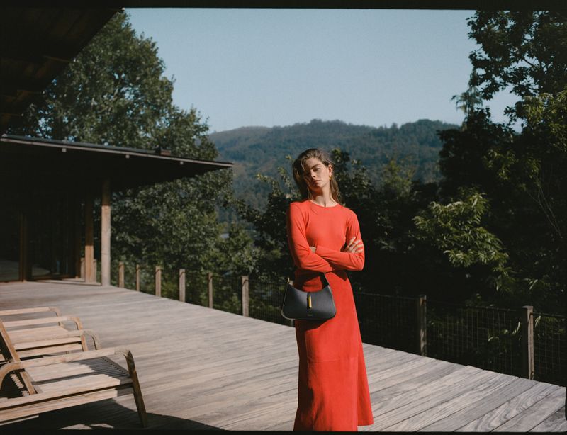 A woman in a long red dress stands on a wooden porch with a gray handbag in front of a background of trees and distant mountains.