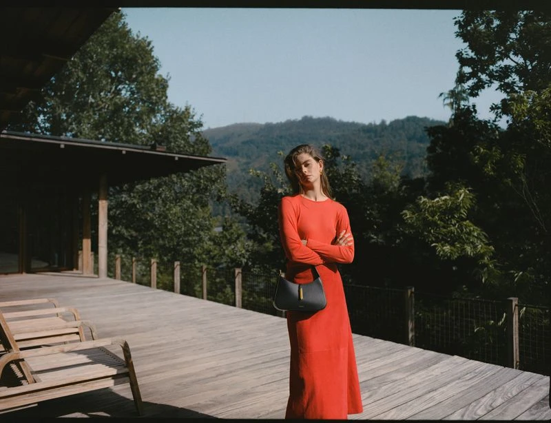 A woman in a long red dress stands on a wooden porch with a gray handbag in front of a background of trees and distant mountains.