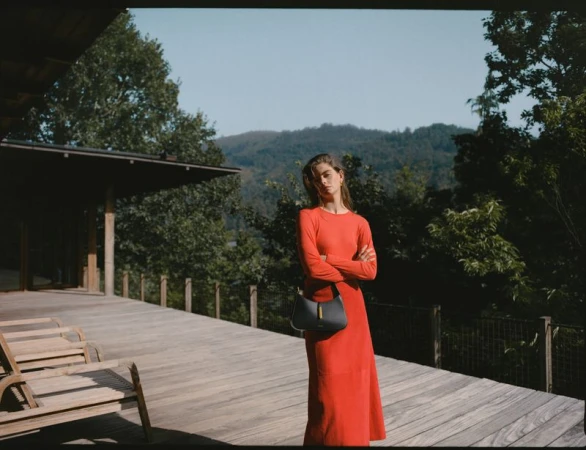 A woman in a long red dress stands on a wooden porch with a gray handbag in front of a background of trees and distant mountains.