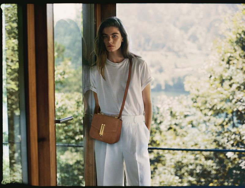 Model Merlijne Schorren in a white t-shirt and white trousers wearing a tan leather crossbody bag by a doorway.