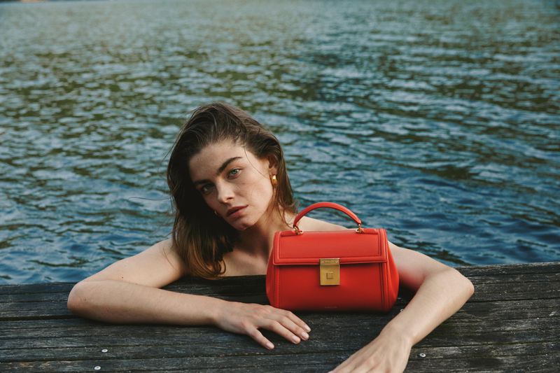 Adult woman poses with a red leather handbag on a wood dock next to rippling water in an eye-level medium shot with natural lighting.