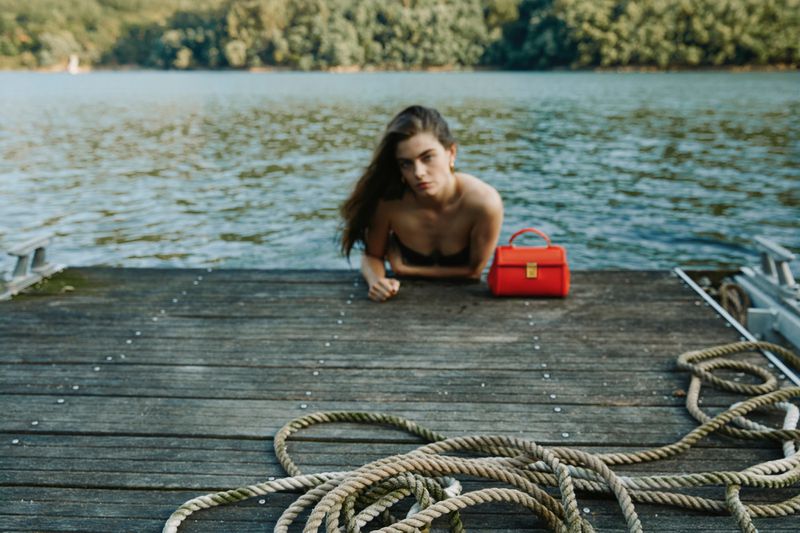Brunette woman on a wooden dock, red handbag, lake background. Outdoors, summertime, natural light, medium shot, with rope in foreground.