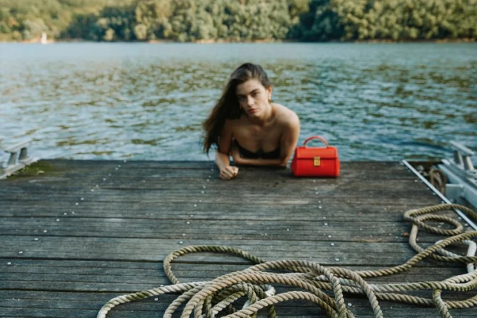 Brunette woman on a wooden dock, red handbag, lake background. Outdoors, summertime, natural light, medium shot, with rope in foreground.