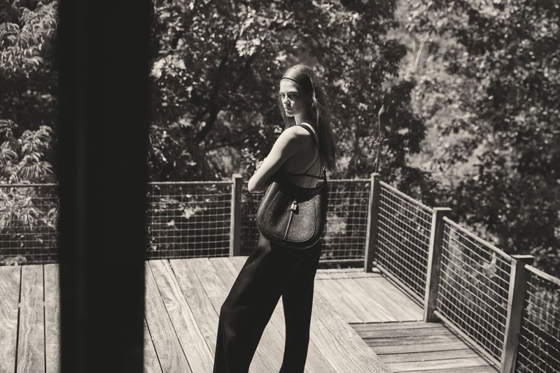 Monochrome shot of a woman with a purse posing on a wood deck, wearing black pants and a strappy top with trees in the background.