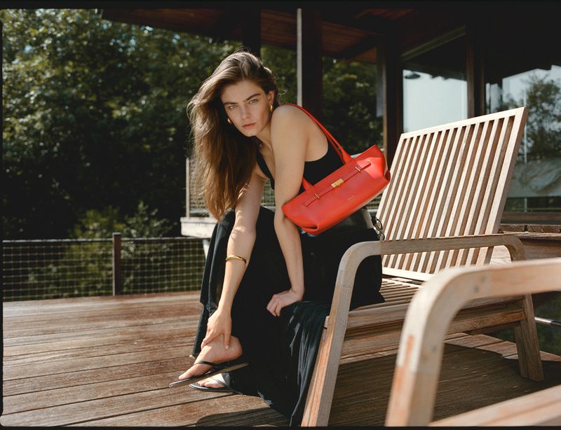 Woman in black dress sits on wooden chair with red leather purse on outdoor deck with green trees, photographed in natural light.