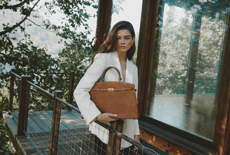 Adult woman in a white blazer poses outdoors, holding a brown suede handbag with gold hardware, standing on a wooden platform.