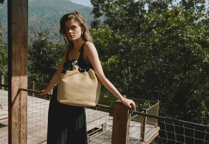 Fashionable woman poses on a wooden balcony with a woven tote bag against a backdrop of lush green trees and rolling mountains.