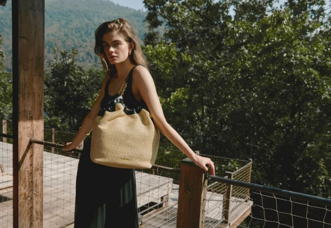 Fashionable woman poses on a wooden balcony with a woven tote bag against a backdrop of lush green trees and rolling mountains.