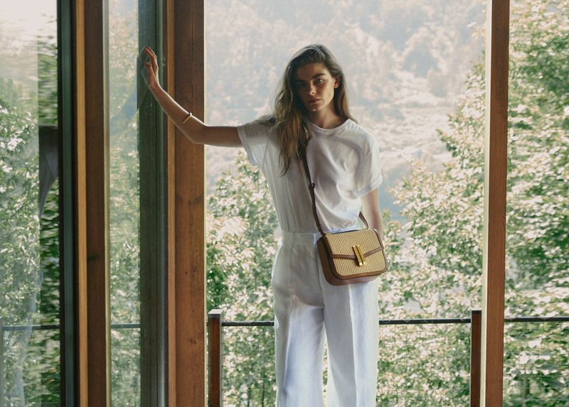 A fashionable woman in a white outfit with a handbag stands near a window, with a scenic view of trees and sky in the background.