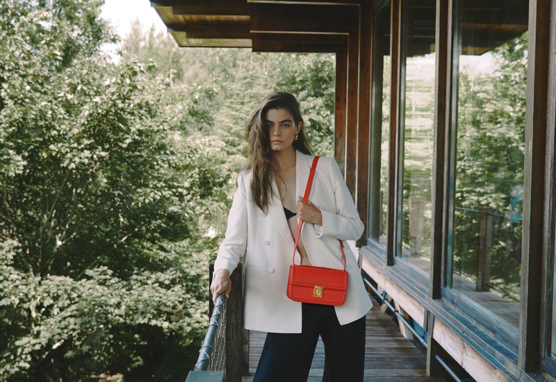 A brunette adult female poses on a wooden porch with a bright orange cross-body purse, wearing a white blazer and black pants.