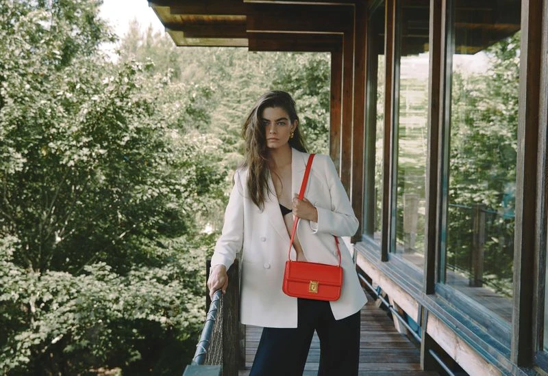 A brunette adult female poses on a wooden porch with a bright orange cross-body purse, wearing a white blazer and black pants.
