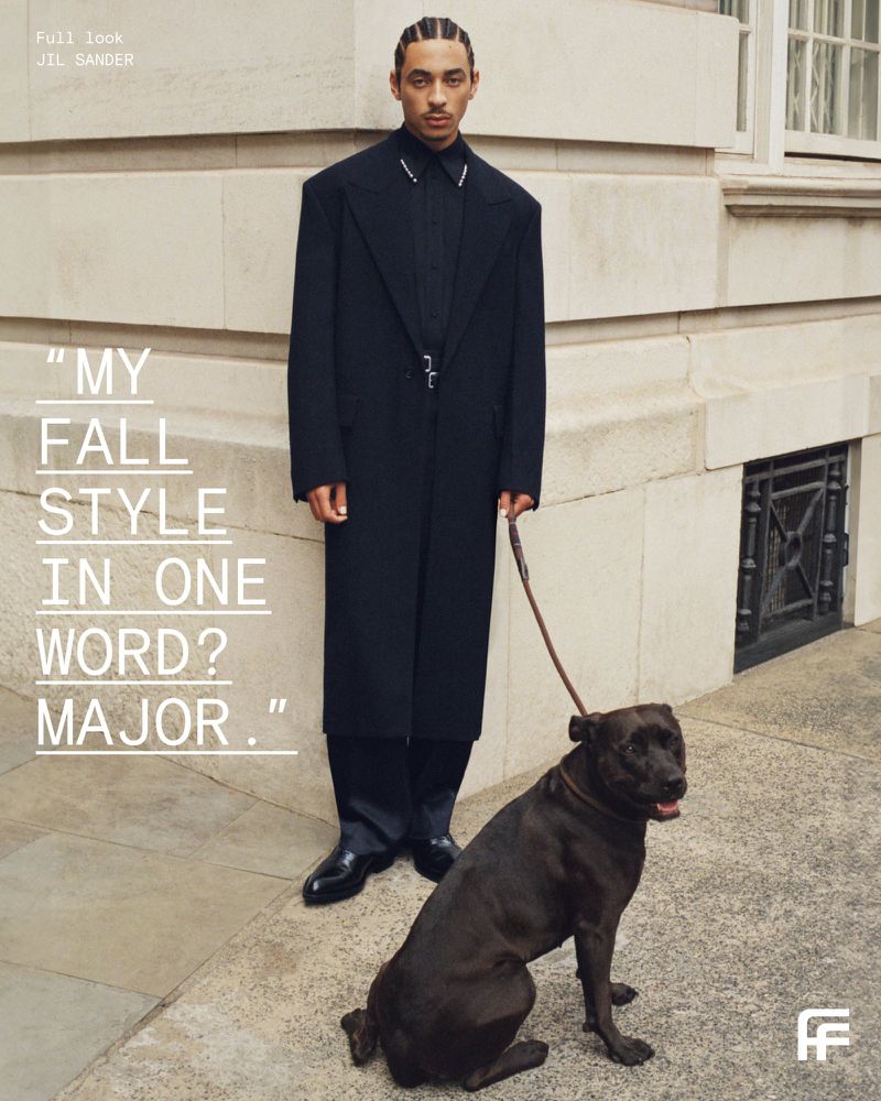 A tall male model and a dog pose on a sidewalk in front of a stone building wearing a long black coat, and black shoes.