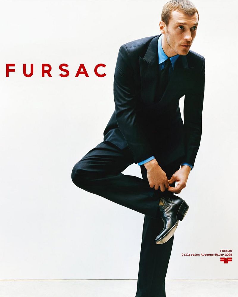 Man in black suit poses adjusting a black boot against a white background with FURSAC logo. Fashion-forward photography.