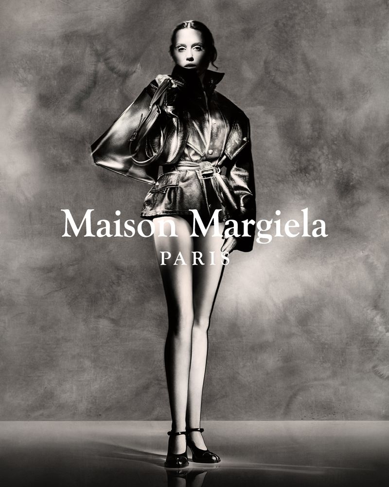 Fashion editorial shot of a slender woman in a black leather jacket and black heels with an abstract grey backdrop and Maison Margiela logo.