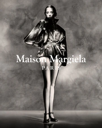 Fashion editorial shot of a slender woman in a black leather jacket and black heels with an abstract grey backdrop and Maison Margiela logo.