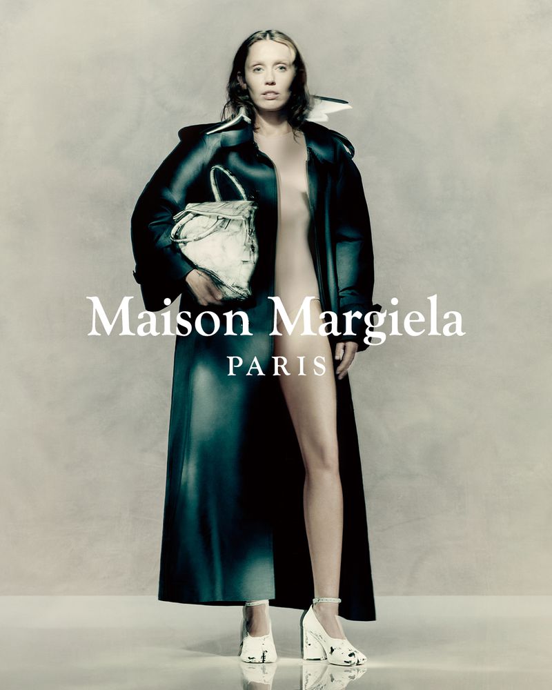 A woman poses in a black coat with a white bag and heels, standing against a neutral backdrop, with brand text overlayed.