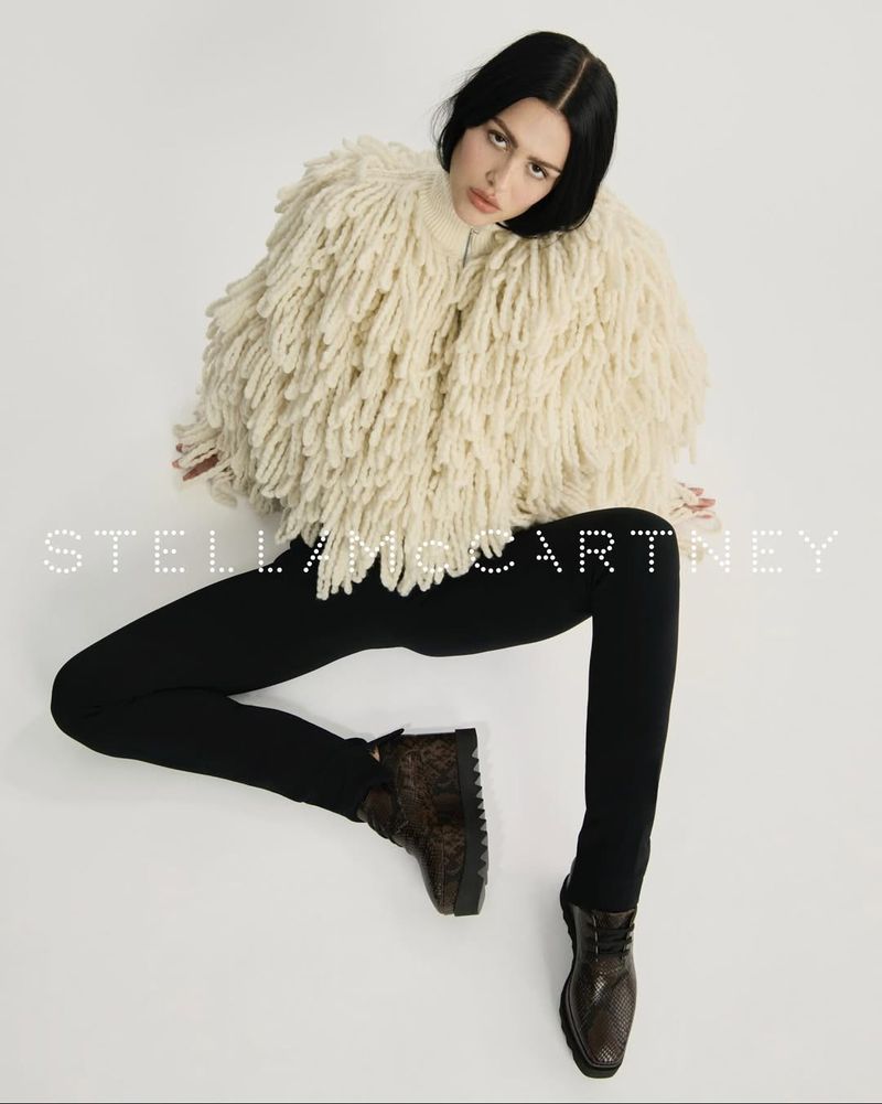 Fashion shot of a young woman with dark hair wearing a shaggy, cream-colored jacket, black pants and dark shoes, sitting against a white backdrop.
