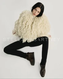 Model wearing a textured cream jacket and black trousers with patterned platform shoes against a white backdrop.