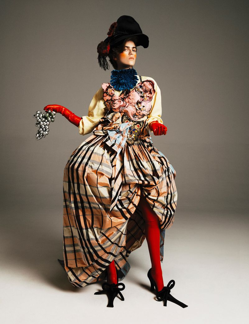 Elegant woman in a unique plaid dress and black hat poses in a studio with silver grapes and red stockings and gloves. Fashion shoot.