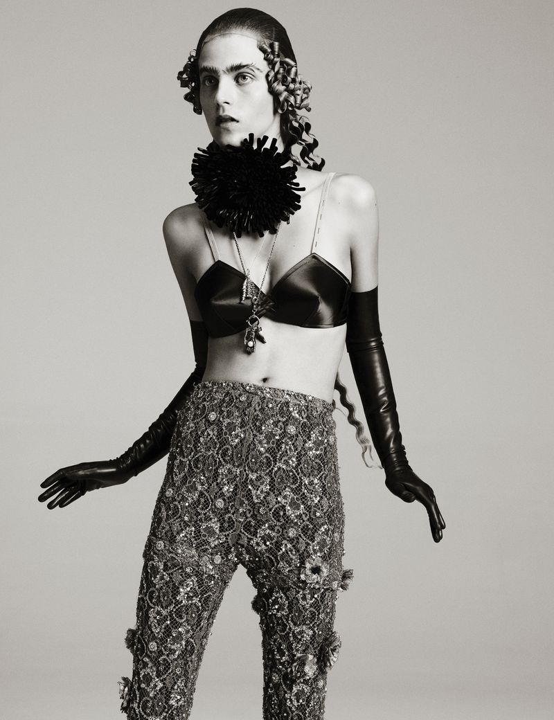 Person in an avant-garde fashion pose wearing a black bra top, beaded pants, long gloves, and a statement necklace on a neutral background.