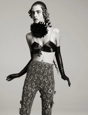 Person in an avant-garde fashion pose wearing a black bra top, beaded pants, long gloves, and a statement necklace on a neutral background.