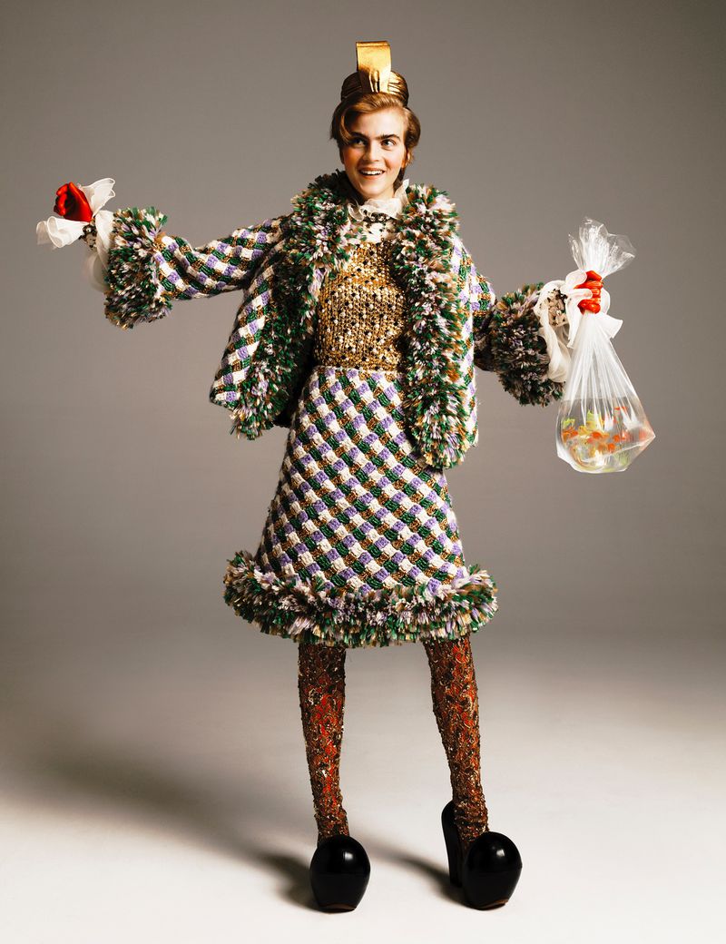 Woman in a tweed jacket and skirt, wearing a gold sequin top and a gold crown, holds bags with red flowers and goldfish against gray backdrop.