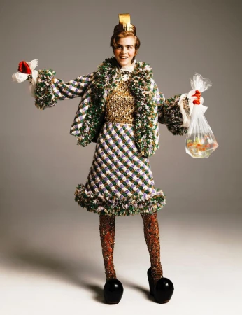 Woman in a tweed jacket and skirt, wearing a gold sequin top and a gold crown, holds bags with red flowers and goldfish against gray backdrop.