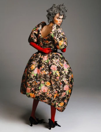 Model wearing a voluminous floral balloon dress, red gloves, and a textured silver headpiece against a grey studio backdrop.