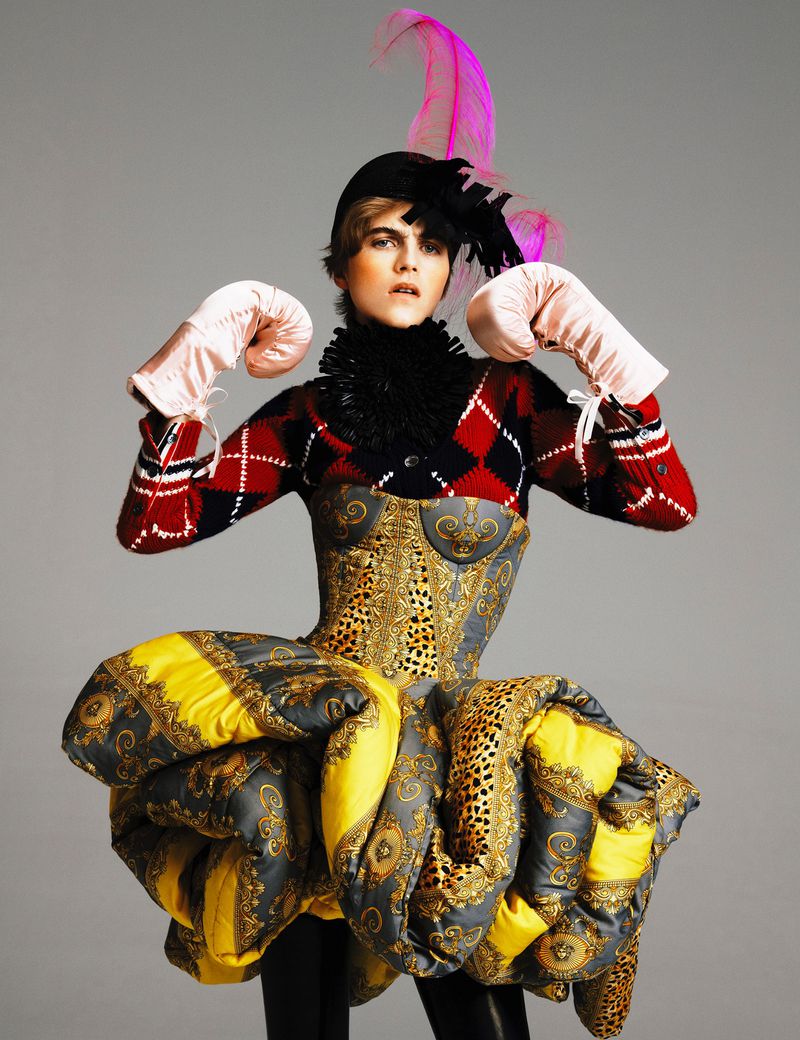 A female fashion model poses in a striking outfit featuring a corset, puffy skirt, boxing gloves, and feathered hat against a grey background.