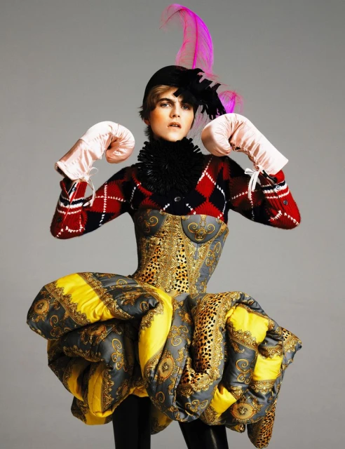 A female fashion model poses in a striking outfit featuring a corset, puffy skirt, boxing gloves, and feathered hat against a grey background.