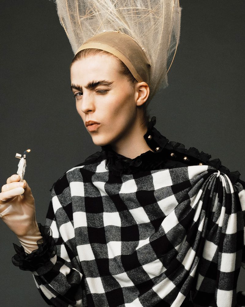 Close-up of a person with dramatic makeup and a large checkered shirt, holding a small figurine, against a dark gray studio backdrop.