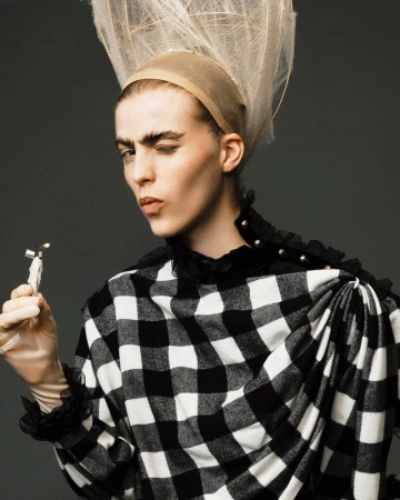 Close-up of a person with dramatic makeup and a large checkered shirt, holding a small figurine, against a dark gray studio backdrop.