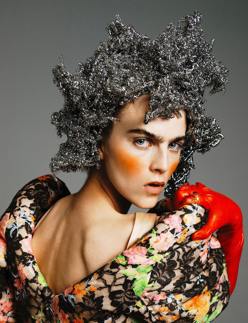 High-fashion portrait of a person with pale skin, wearing an intricate silvery headpiece and a floral dress against a grey backdrop.