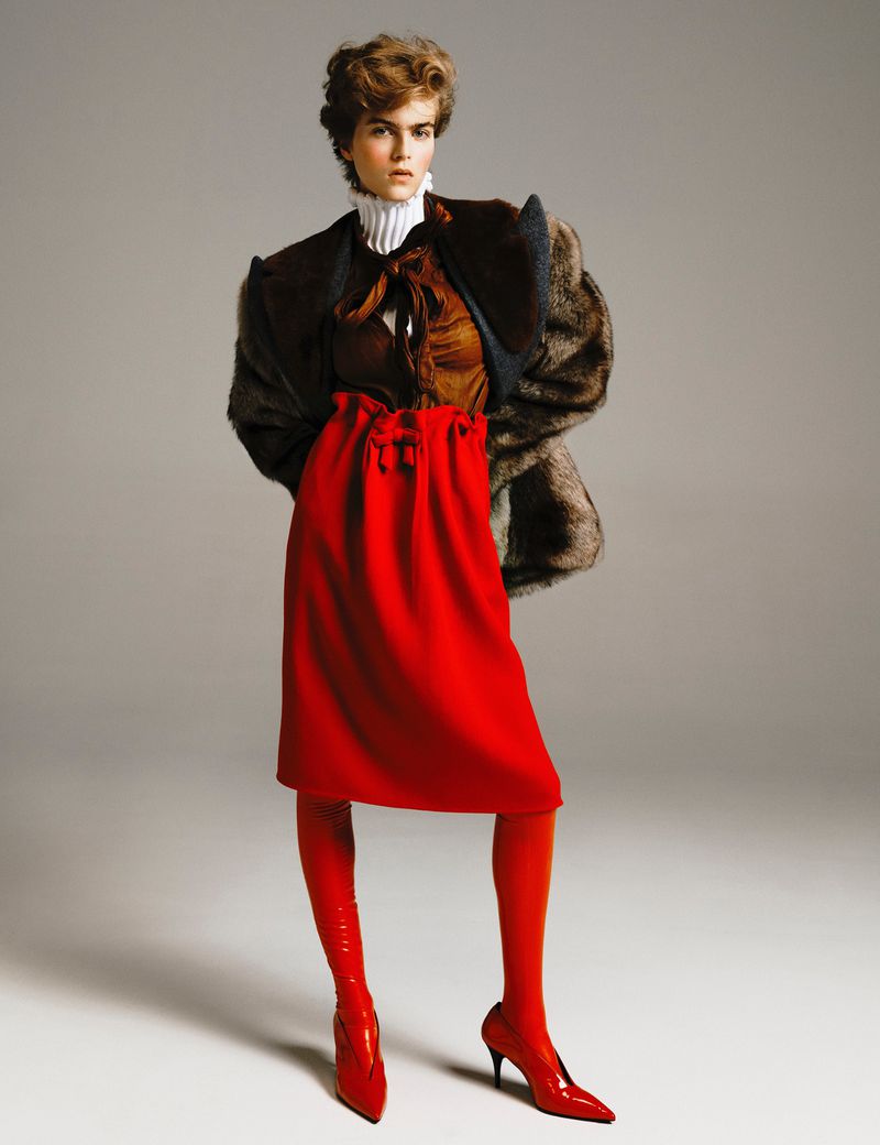 A woman poses in a brown top, red skirt, fur coat, and matching boots against a neutral background in a fashion editorial studio shot.